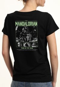 Svart t-shirt med ett grafiskt tryck av "The Mandalorian" på baksidan, med grön text och bilder av karaktärer i ett fyrkantigt format.