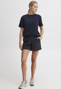Marineblå bomulls t-skjorte med brystlomme, kombinert med mørkegrå teksturerte shorts. Modellen har på seg nøytrale joggesko og står mot en enkel bakgrunn.