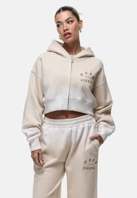 Haut à capuche beige raccourci avec une fermeture éclair à l'avant et une grande capuche. Présente une texture douce et un motif imprimé. Associé à un jogging décontracté assorti.