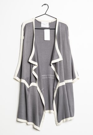 Cardigan gris ouvert devant avec bordure blanche, suspendu sur un cintre noir sur fond blanc.