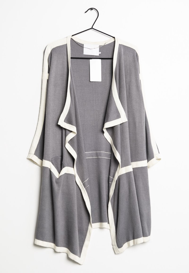 Cardigan gris ouvert devant avec bordure blanche, suspendu sur un cintre noir sur fond blanc.