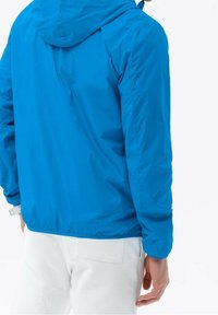 Blå letvægtsjakke med hætte, der har en glat tekstur og elastisk kant. Bruges over hvide bukser og har et afslappet design.