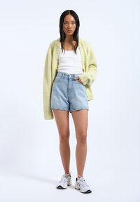 Gilet tricoté jaune clair, débardeur blanc, short en denim bleu clair taille haute et baskets blanches sur un fond uni.