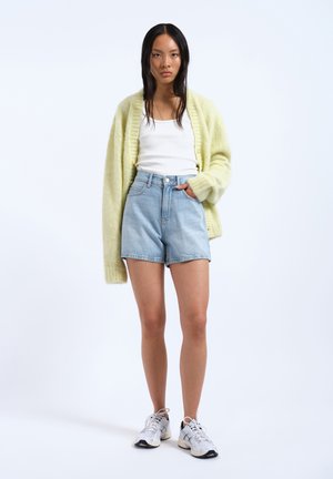 Pletený světle žlutý svetr, bílý top na ramínka, světlé modré džíny s vysokým pasem a bílé sportovní boty na jednobarevném pozadí.