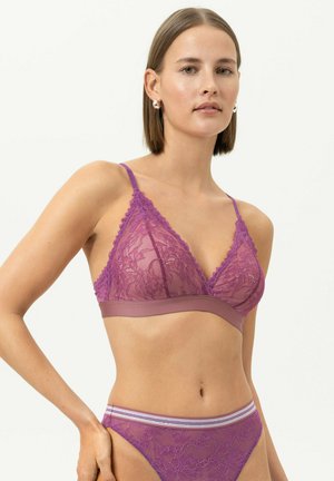 Femme aux cheveux bruns lisses portant un bralette en dentelle violette et une culotte assortie en dentelle avec une ceinture rayée sur un fond uni.