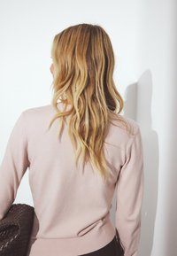 Gaiši rozā džemperis ar mīkstu tekstūru, garām piedurknēm un ribota apakšmalu. Individuāls tur tumši brūnu austu rokassomu.