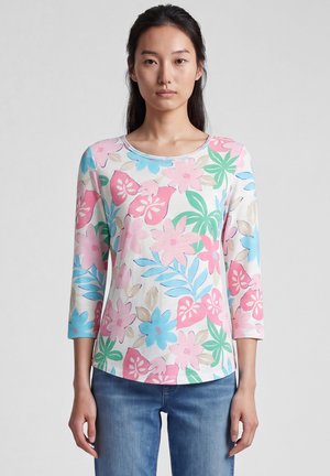 Frau trägt ein langärmeliges weißes Hemd mit pastellrosa, blauem und grünem Blumenmuster und blaue Jeans, steht vor einem schlichten Hintergrund.