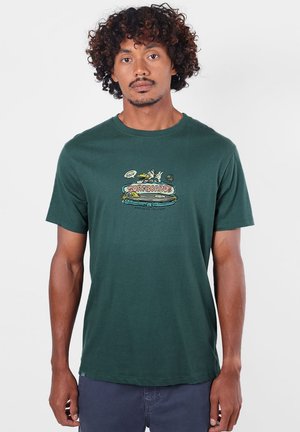 Homme aux cheveux bouclés portant un t-shirt vert foncé avec un motif planche de surf colorée et le texte "Surfboards", debout devant un fond uni.