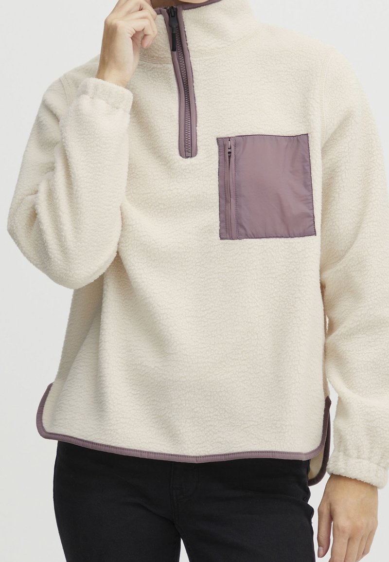 Pull polaire beige clair avec bordure mauve, zip au quart et poche poitrine zippée porté par une personne ajustant le zip.