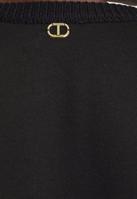 Pull noir avec un col en maille et un accent en métal doré portant le nom de la marque "TWINSET" au dos. Texture lisse.