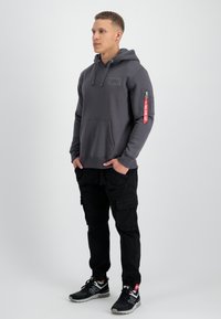 Sudadera con capucha gris, con bolsillo frontal, bolsillo con cremallera en el brazo y detalle de etiqueta roja; combinada con pantalones cargo negros y zapatillas negras.