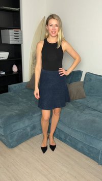 Mujer con cabello largo y rubio, vistiendo una blusa negra sin mangas, falda azul marino y zapatos negros puntiagudos, de pie en una sala de estar cerca de un sofá azul.