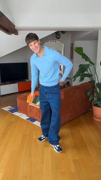 Νεαρός άνδρας που χαμογελά, φορώντας μπλε πουκάμισο με μακριά μανίκια και φαρδιά τζιν, στέκεται σε έναν μοντέρνο καθιστικό με πορτοκαλί καναπέ και μεγάλο φυτευτό φυτό.