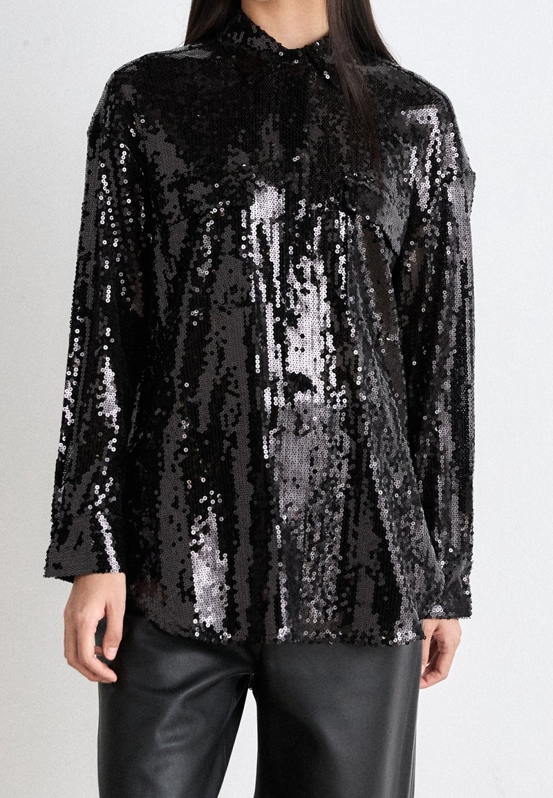 Black sequin shirt with long sleeves, classic collar, and a loose fit. Textured, reflective surface with a shiny finish, paired with leather pants.