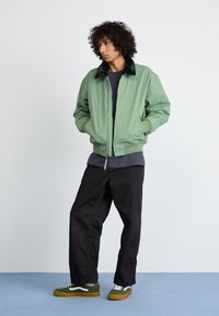 Grøn bomberjakke med sort faux fur-krave, ribbede manchetter og kant, båret over en navy t-shirt, parret med sorte bukser og grønne sneakers.