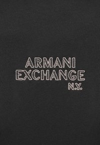 Melnā audumā ar izšūtu "ARMANI EXCHANGE N.Y." gaiši rozā krāsā, ietverot treknus bloku burti uz gludas, strukturētas virsmas.
