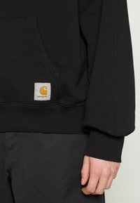 Felpa nera con tasca frontale, polsini a coste e un’etichetta quadrata con un logo giallo. Tessuto morbido con una superficie liscia.