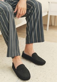 Svarta, vävda slip-on-skor med ett mjukt innerfoder, använda med grå randiga byxor, på en ljus texturerad matta. Den anmärkningsvärda designen är minimalistisk.