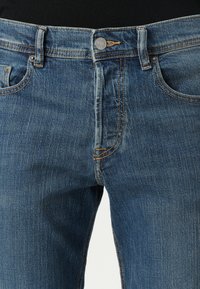 Jeans de mezclilla en azul medio, con corte clásico, costuras visibles, cierre de metal y bolsillos frontales con un ligero desgaste.
