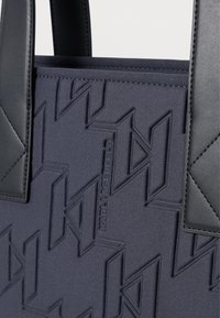 Mala tote azul marinho com padrão texturizado, apresentando detalhes em couro preto liso e alças robustas, desenhada com um detalhe de logotipo em relevo.