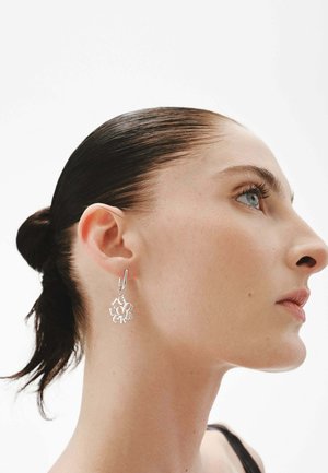 Pendientes de plata con un diseño moderno entrelazado, que presenta formas de letras variadas, colgando de un pequeño aro, llevados en la oreja de un modelo.