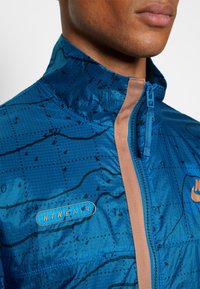 Sininen Nike-takki, jossa on geometrinen kuvio, korkea kaulus, beige vastavärinen vetoketju ja "NIKE AIR" -logo rinnassa.