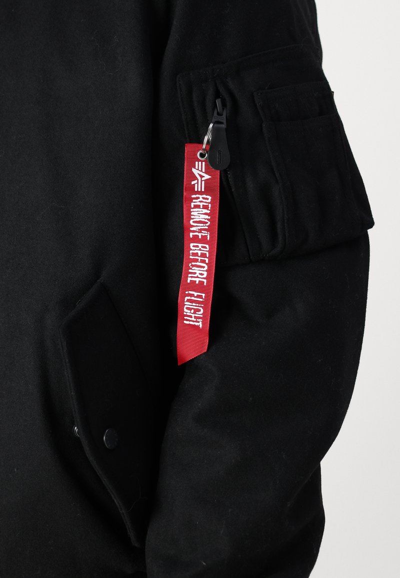 Schwarze Bomberjacke mit strukturierter Oberfläche, ausgestattet mit einem roten Stoffetikett mit der Aufschrift "VOR FLUG ENTFERNEN" und einer externen Tasche am Ärmel.