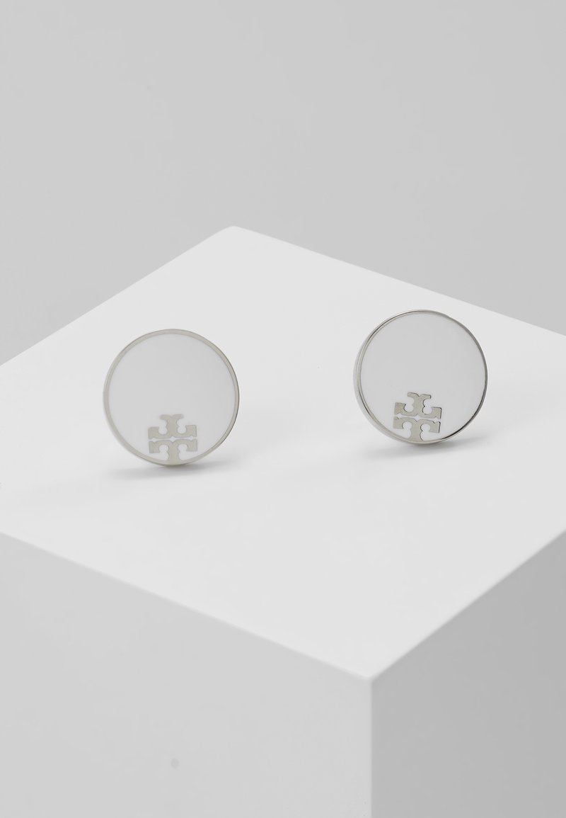 Boucles d’oreilles rondes en argent avec une surface lisse et réfléchissante et un petit logo en forme de croix près du bord inférieur sur un piédestal blanc.