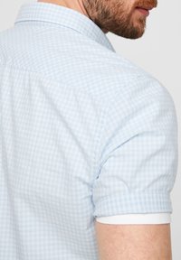 Světle modrá krátkosrstá kostkovaná košile s klasickým límečkem, ohrnutými rukávy s bílými akcenty a texturovanou tkaninou.
