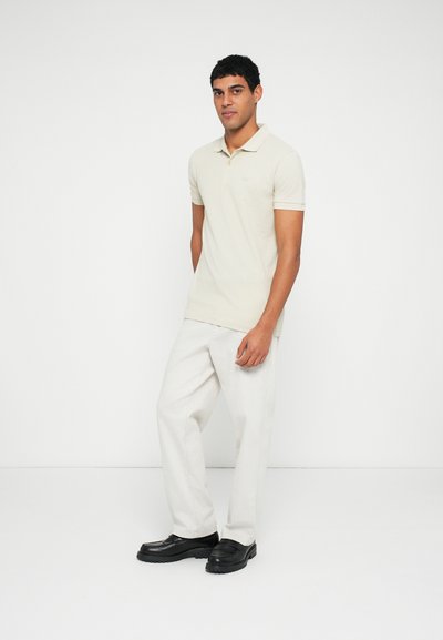 Polo beige clair à manches courtes avec un logo discret, associé à un pantalon large couleur crème et des mocassins noirs.