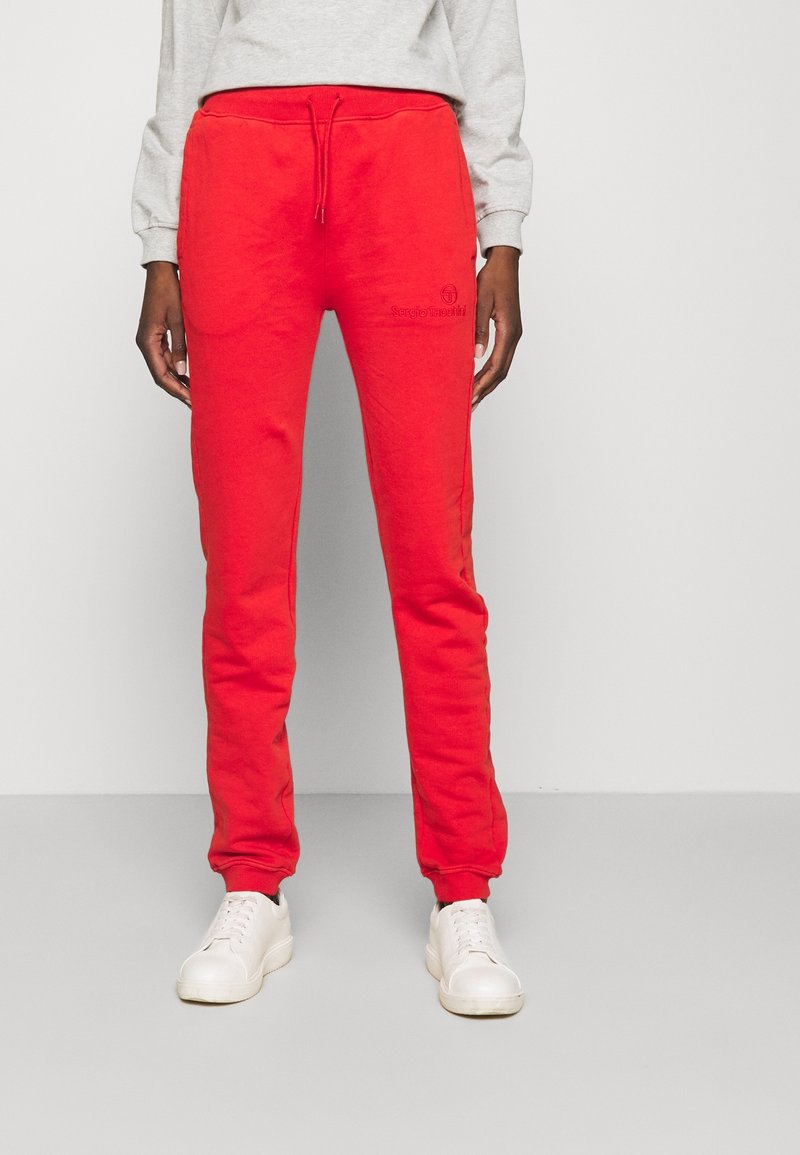 Red sweatpants with an elastic waistband and drawstring, featuring a logo embroidery. The fabric appears soft and textured, paired with white sneakers.