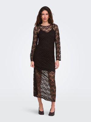 Femme portant une longue robe noire en dentelle avec manches transparentes et superposition de jupe, associée à des talons noirs, debout devant un fond uni.