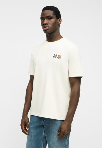 Νεαρός άντρας που φοράει λευκό μπλουζάκι με μικρές κεντημένες κεφαλές λύκου και αλεπούς, συνδυασμένο με μπλε τζιν, στέκεται μπροστά σε απλό φόντο.