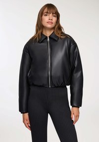 Kobieta w czarnej kurtce z zamkiem błyskawicznym z ekoskóry z kołnierzem oraz czarnych dopasowanych spodniach w prążki, stojąca na jednolitym tle.