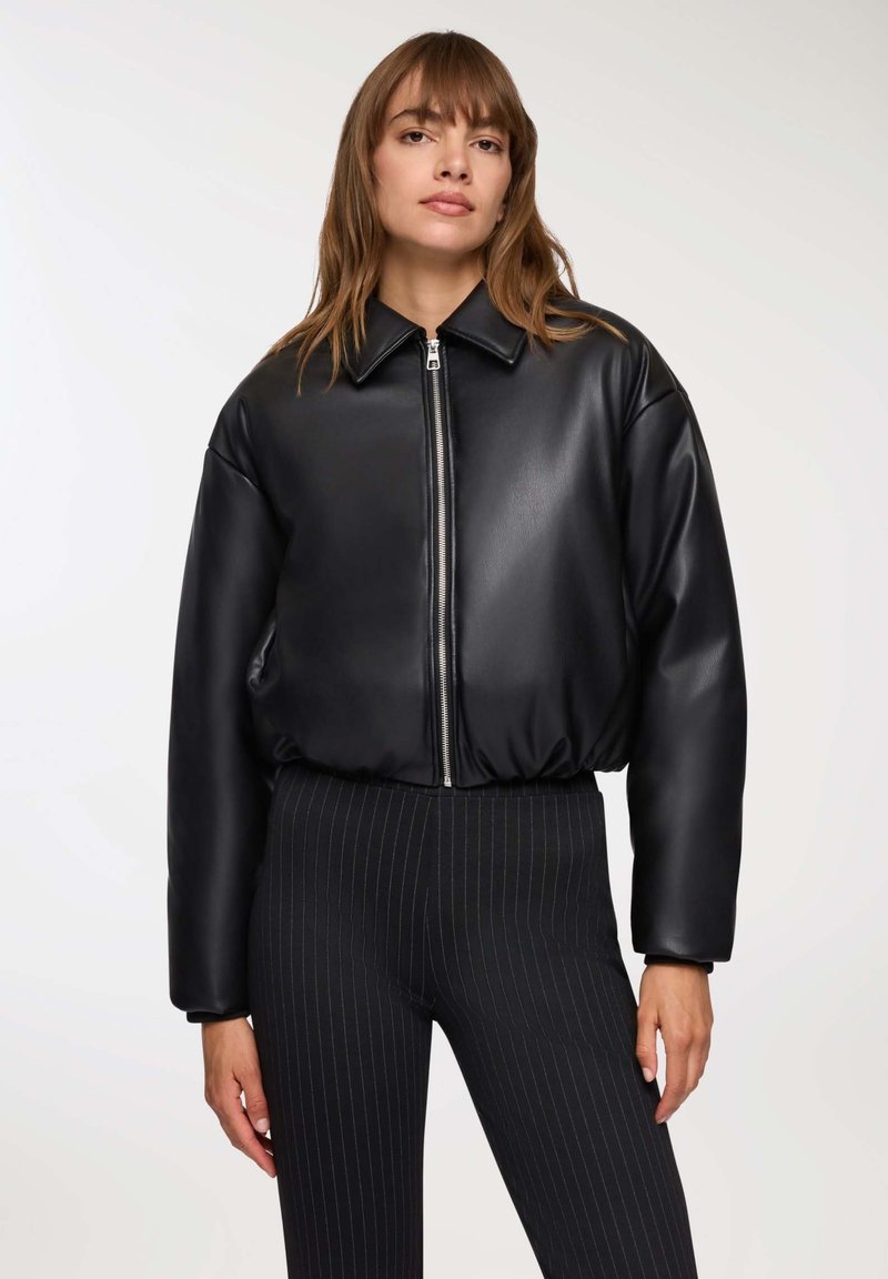 Kobieta w czarnej kurtce z zamkiem błyskawicznym z ekoskóry z kołnierzem oraz czarnych dopasowanych spodniach w prążki, stojąca na jednolitym tle.