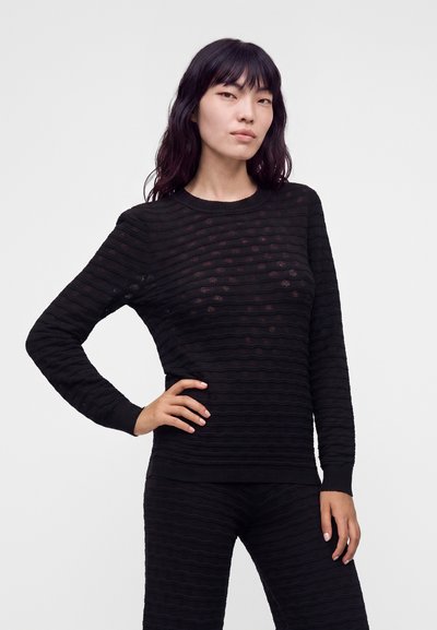 Femme aux longs cheveux foncés portant un haut noir texturé à manches longues et un pantalon assorti, debout avec une main sur la hanche.