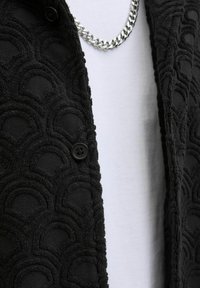 Black textured jacket featuring a wave pattern, with a button detail and a silver chain necklace over a white shirt.