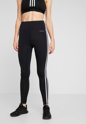 Leggings deportivos negros con franjas laterales blancas, diseño de cintura alta y una textura suave y elástica. Combinados con zapatillas deportivas negras.