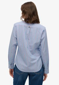 Kvinde med skulderlangt brunt hår iført en blå skjorte med tynde lyserøde lodrette striber og blå jeans, set bagfra.