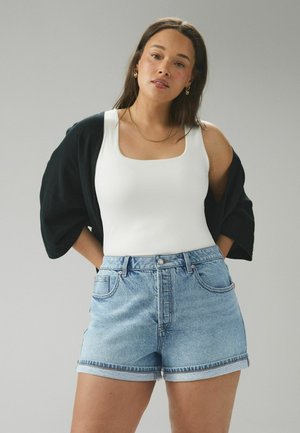 Femeie purtând un top alb, pantaloni scurți din denim deschis la culoare și un cardigan negru așezat pe umeri, stând în fața unui fundal gri neutru.