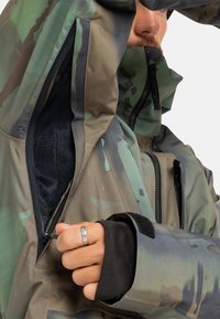 Maskáčová bunda v tlumených zelených a hnědých barvách; obsahuje kapsy na zip, síťovou podšívku a nastavitelné manžety se suchými zipy.