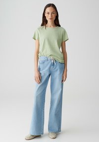 T-shirt verde chiaro a maniche corte abbinato a jeans larghi azzurri. I jeans presentano una vita con coulisse e una texture leggermente sbiadita.