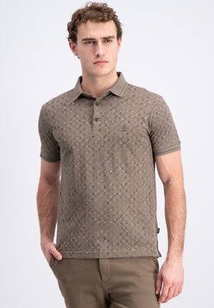 Jonge man met een bruin patroon polohemd en bruine broek, staat met één hand in de zak en kijkt opzij.