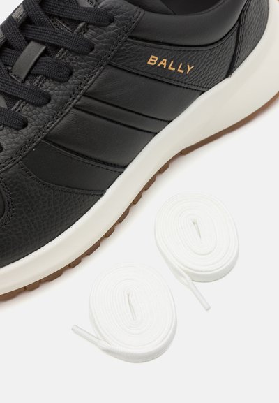 Zapatilla negra de cuero con acabado texturizado, tres franjas horizontales y suela de goma blanca. Dos cordones blancos se encuentran al lado del zapato.