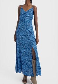 Robe longue bleue à motif tacheté, avec fines bretelles, un détail froncé à l'avant et une fente haute sur la cuisse, associée à des bottes hautes marron clair.