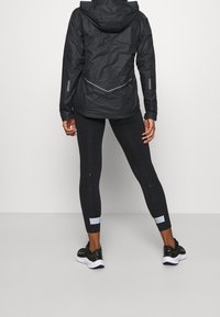 Czarna kurtka z zamkiem błyskawicznym i kapturem, w połączeniu z teksturowanymi czarnymi legginsami z subtelnym wzorem oraz czarnymi butami sportowymi z białymi podeszwami.