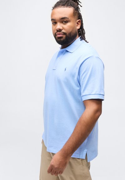 Άνδρας με πλεξούδες, φορώντας ανοιχτό γαλάζιο πόλο και μπεζ παντελόνι, στέκεται μπροστά σε απλό λευκό φόντο.