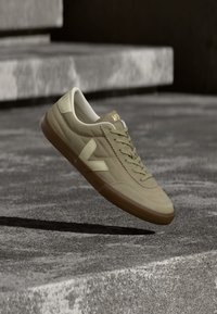 Μπεζ δερμάτινο αθλητικό παπούτσι με ανοιχτόχρωμη σόλα και λογότυπο «V», αιωρούμενο πάνω από γκρί τσιμεντένιο έδαφος και σκαλοπάτια σε έναν μινιμαλιστικό εξωτερικό χώρο.