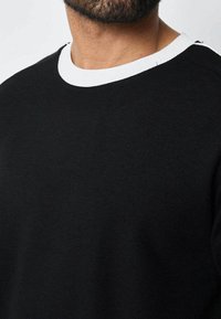 Pull noir en tissu texturé avec un col rond côtelé blanc. Le design est uni et ne comporte pas de motifs ou d'accents supplémentaires.