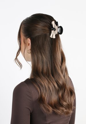 Una fermaglietta per capelli con una base nera e un accento in nastro crema. Progettata a forma di fiocco, tiene indietro capelli castani mossi di media lunghezza.